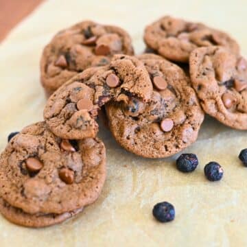 Blueberry Chocolate Chip Cookies rest on parchment paper, with some cookies stacked and one featuring a tempting bite. Scattered blueberries and rich chocolate chips make these treats irresistible.