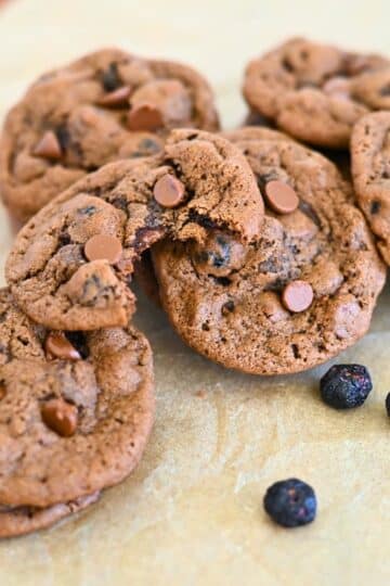 Blueberry Chocolate Chip Cookies rest on parchment paper, with some cookies stacked and one featuring a tempting bite. Scattered blueberries and rich chocolate chips make these treats irresistible.