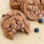 Close-up of blueberry chocolate chip cookies, one with a bite taken, resting on parchment paper with scattered chocolate chips.