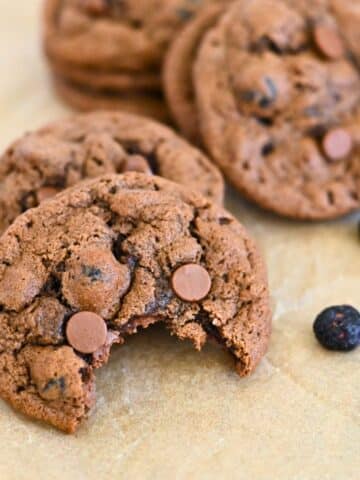 Close-up of blueberry chocolate chip cookies, one with a bite taken, resting on parchment paper with scattered chocolate chips.