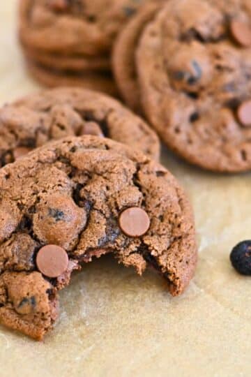 Close-up of blueberry chocolate chip cookies, one with a bite taken, resting on parchment paper with scattered chocolate chips.