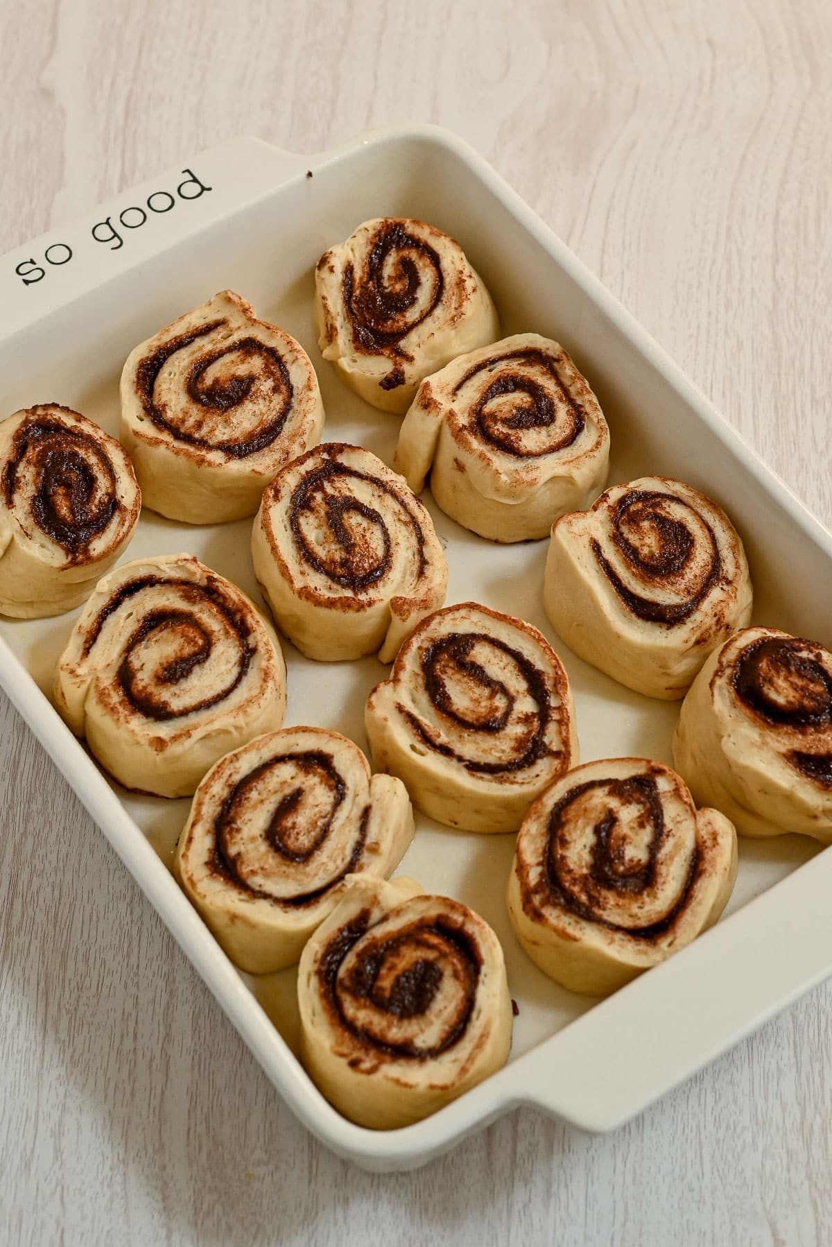 Unbaked chocolate cinnamon rolls in a white rectangular baking dish on a light wood surface.