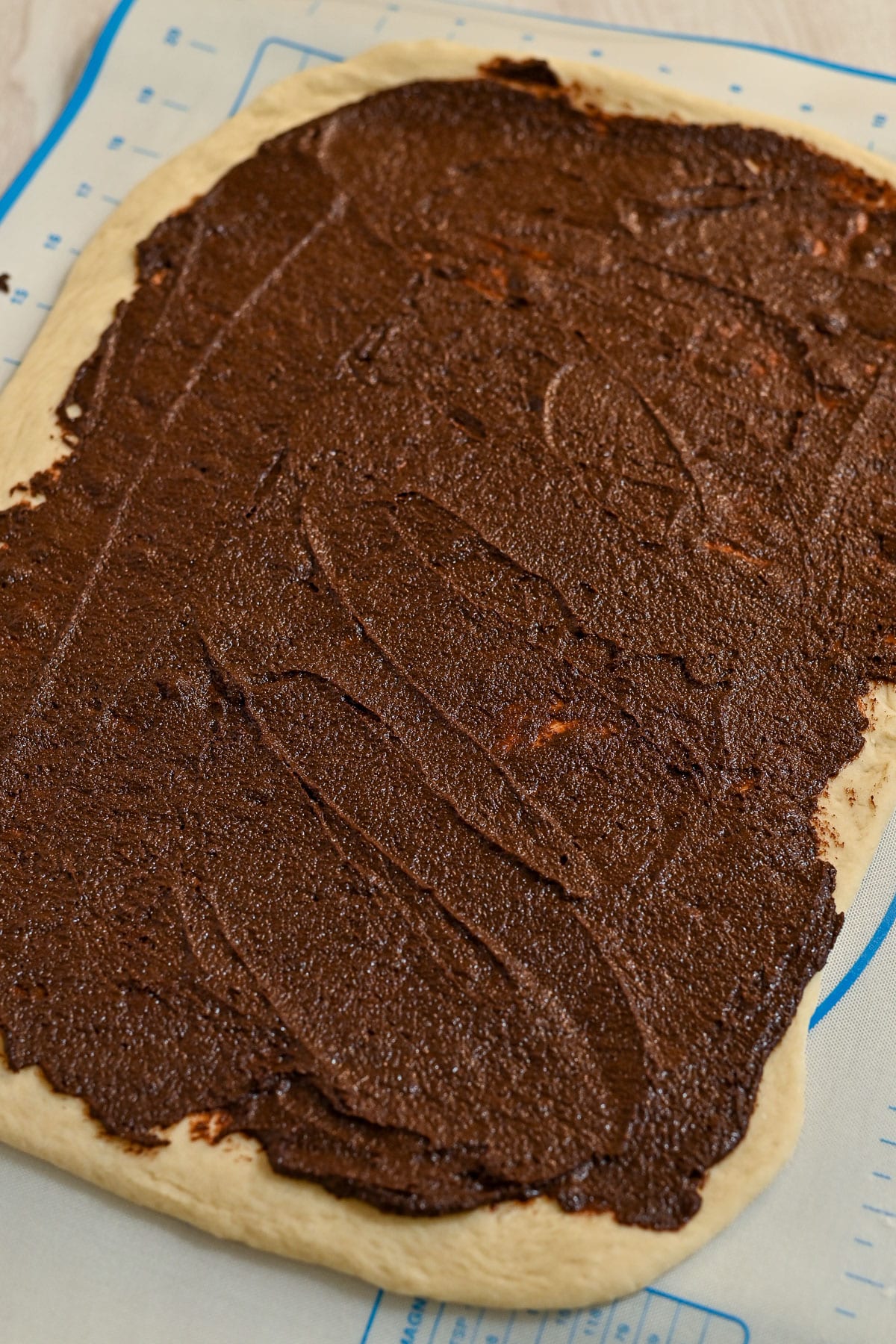 Rectangle of dough with chocolate spread on top, ready to be rolled or baked into delicious chocolate cinnamon rolls, resting on a baking mat.