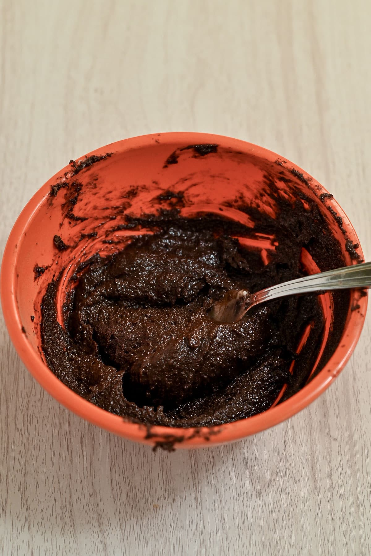 A spoon in a red bowl with thick chocolate batter for cocoa cinnamon rolls rests on a light wooden surface.