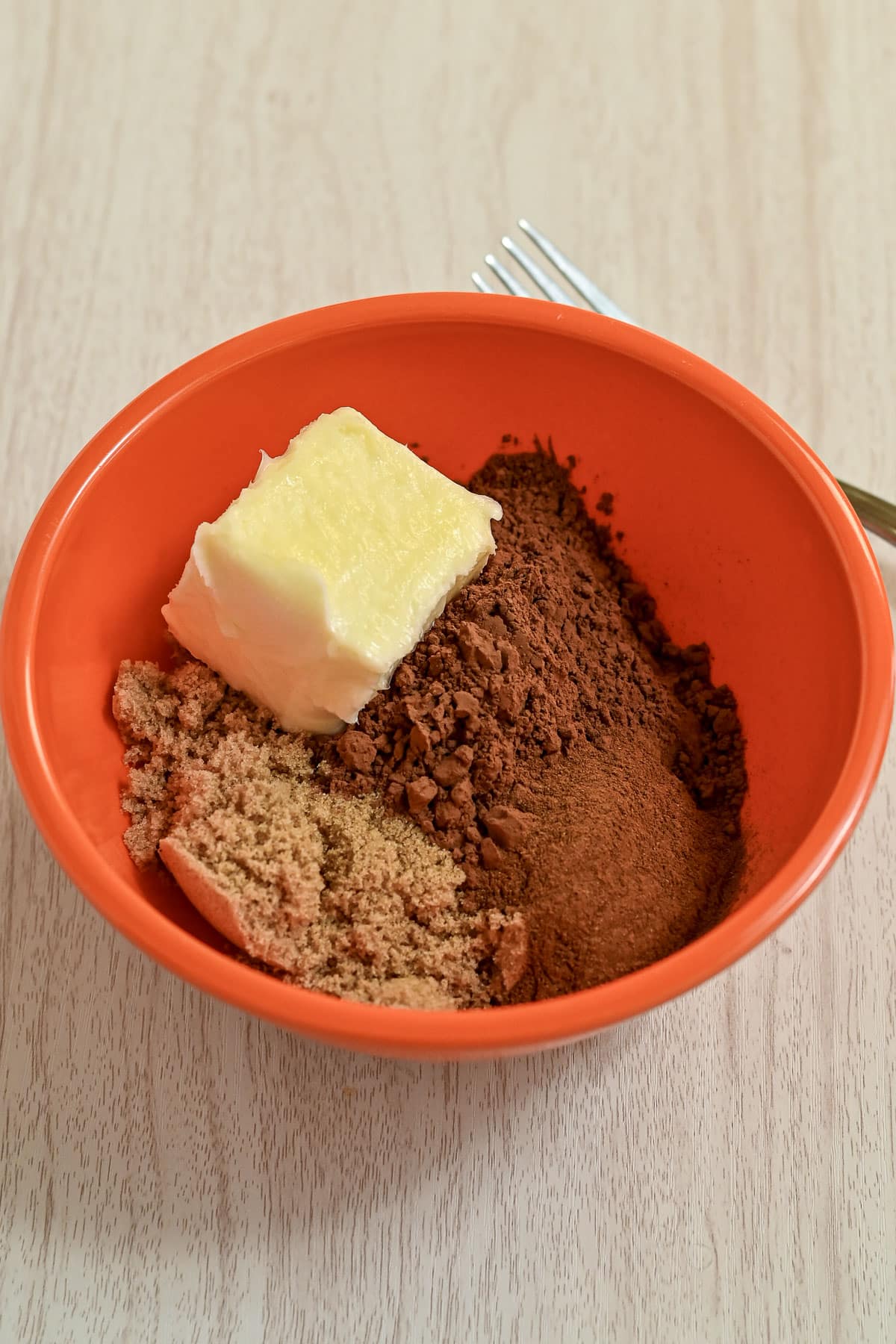 An orange bowl with butter, cocoa powder, brown sugar, and cinnamon sits on a light wooden surface-the perfect beginning for rich and gooey chocolate cinnamon rolls.