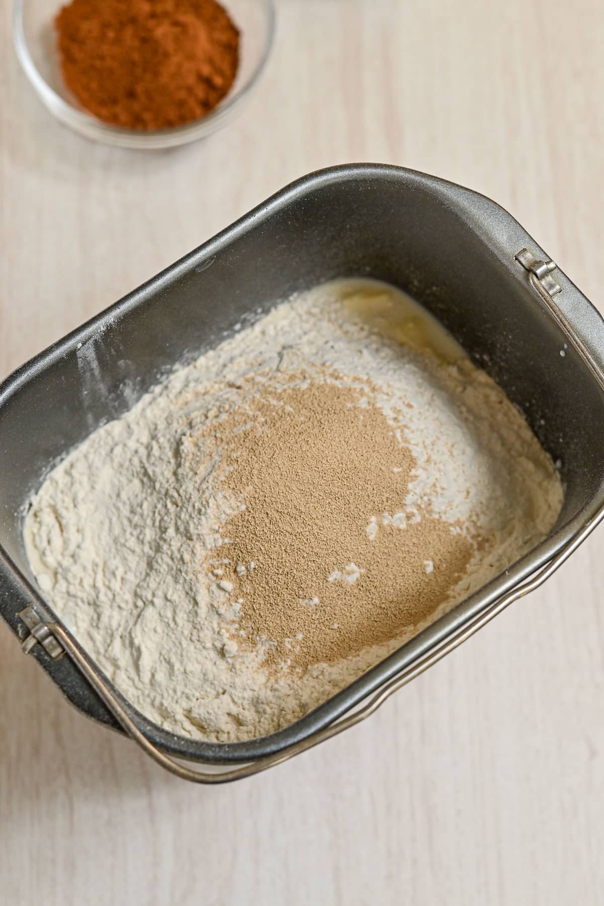 Bread machine pan with flour, yeast, and liquid-ready for making delicious chocolate cinnamon rolls; a small bowl of spices in the background on a light surface.