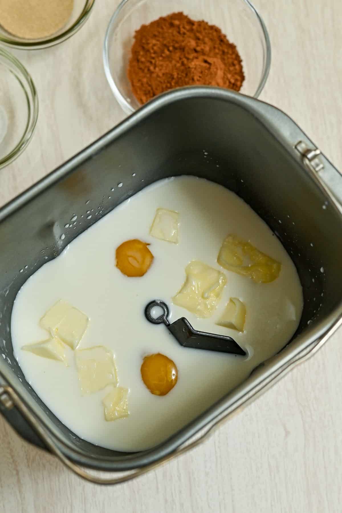 Milk, eggs, and butter in a bread machine pan, with bowls of cocoa and sugar nearby on a light surface-everything you need to start baking delicious cocoa cinnamon rolls.