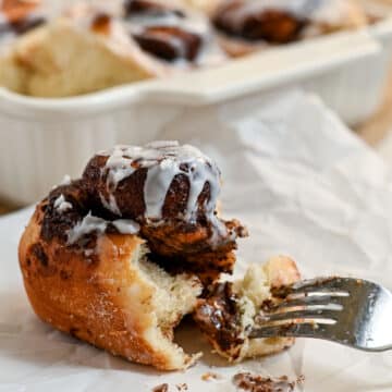 A fork cuts into a gooey chocolate cinnamon roll with white icing, with more cocoa cinnamon rolls in a baking dish in the background.