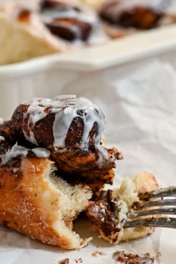 A fork cuts into a gooey chocolate cinnamon roll with white icing, with more cocoa cinnamon rolls in a baking dish in the background.