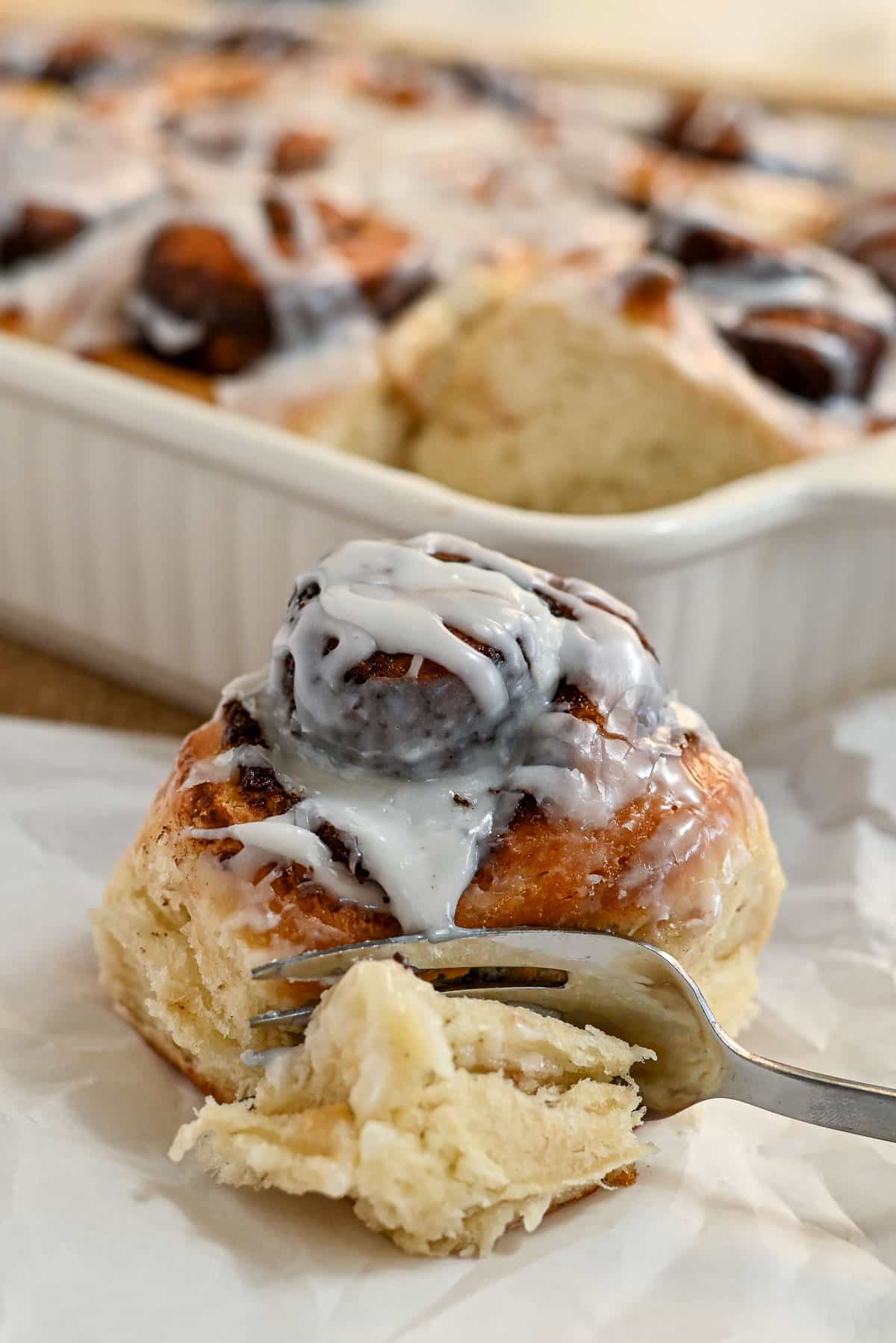 A fork cuts into a cinnamon roll with icing, while a pan of freshly baked cocoa cinnamon rolls sits in the background.
