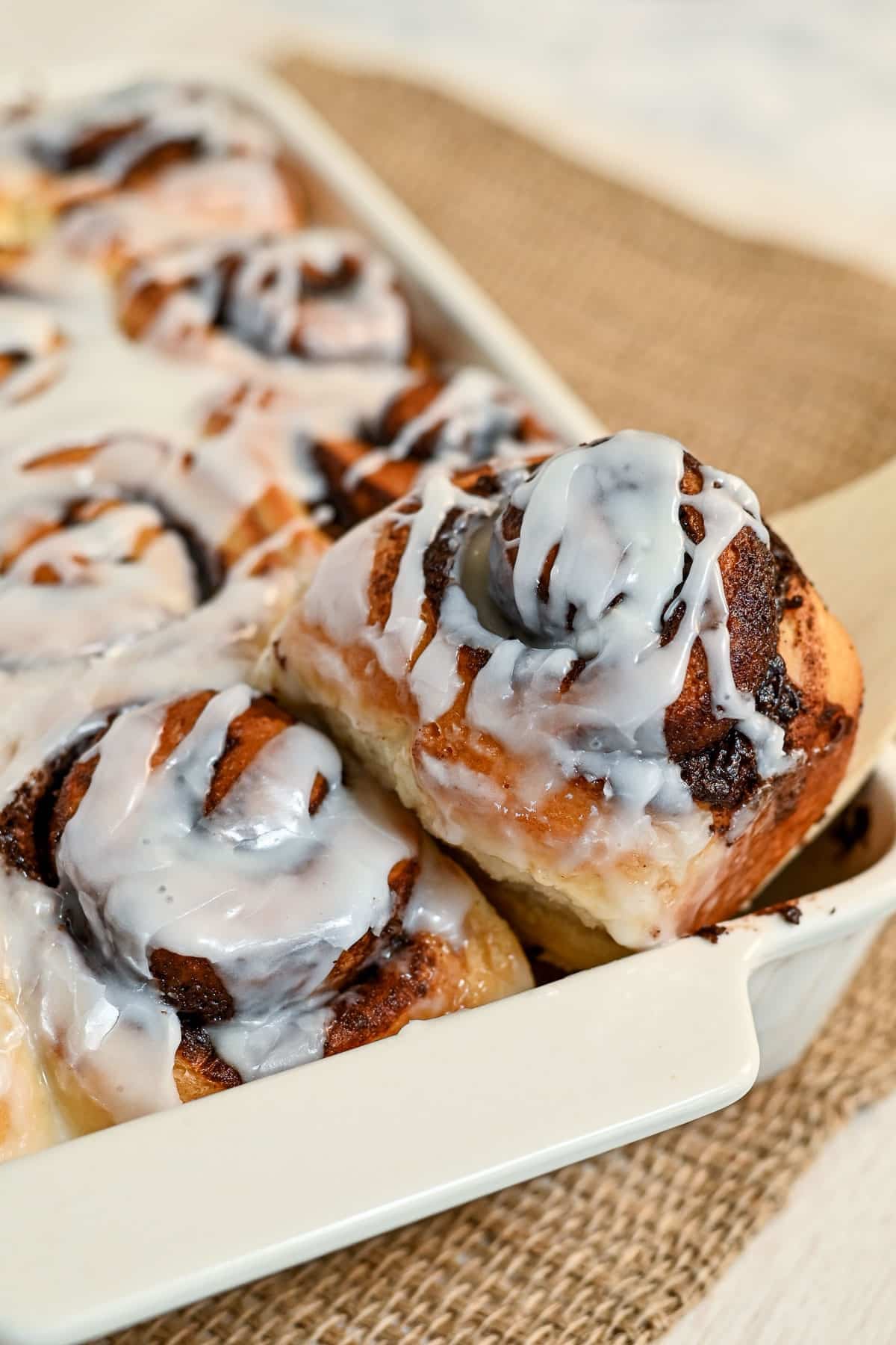 Freshly baked Chocolate Cinnamon Rolls with white icing in a baking dish, one cocoa cinnamon roll is being pulled out.