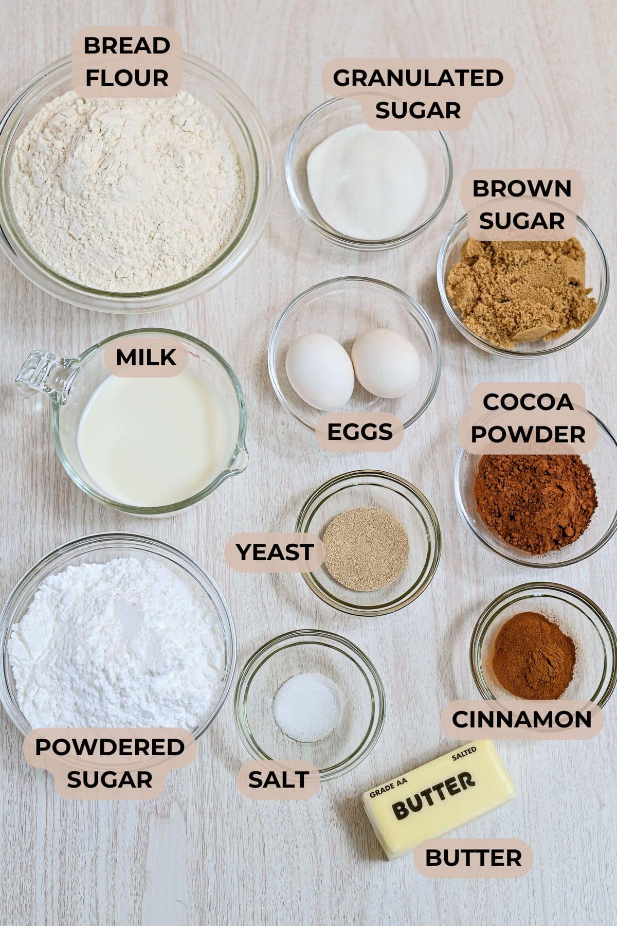 Top-down view of labeled bowls containing baking ingredients on a light surface, including flour, sugars, eggs, and cocoa powder-perfect for making decadent chocolate cinnamon rolls or classic cinnamon rolls.