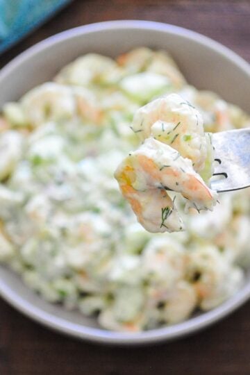 two shrimp on a fork hovering over a gray bowl filled with shrimp salad