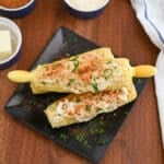 Two ears of Kicked-Up Corn on the Cob, topped with creamy sauce, bbq spice, and chopped herbs, are served on a black plate. Additional condiment bowls and a cloth napkin are in the background.