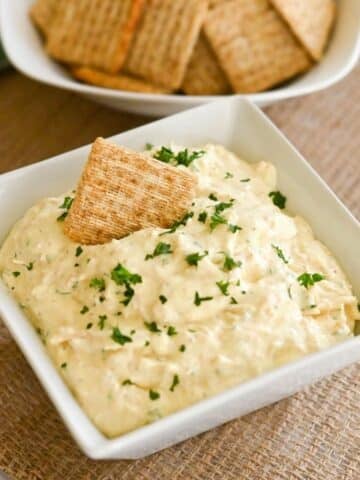 A square bowl of creamy chicken dip topped with parsley sits invitingly, surrounded by crispy crackers on a textured mat, with more crackers in the background.