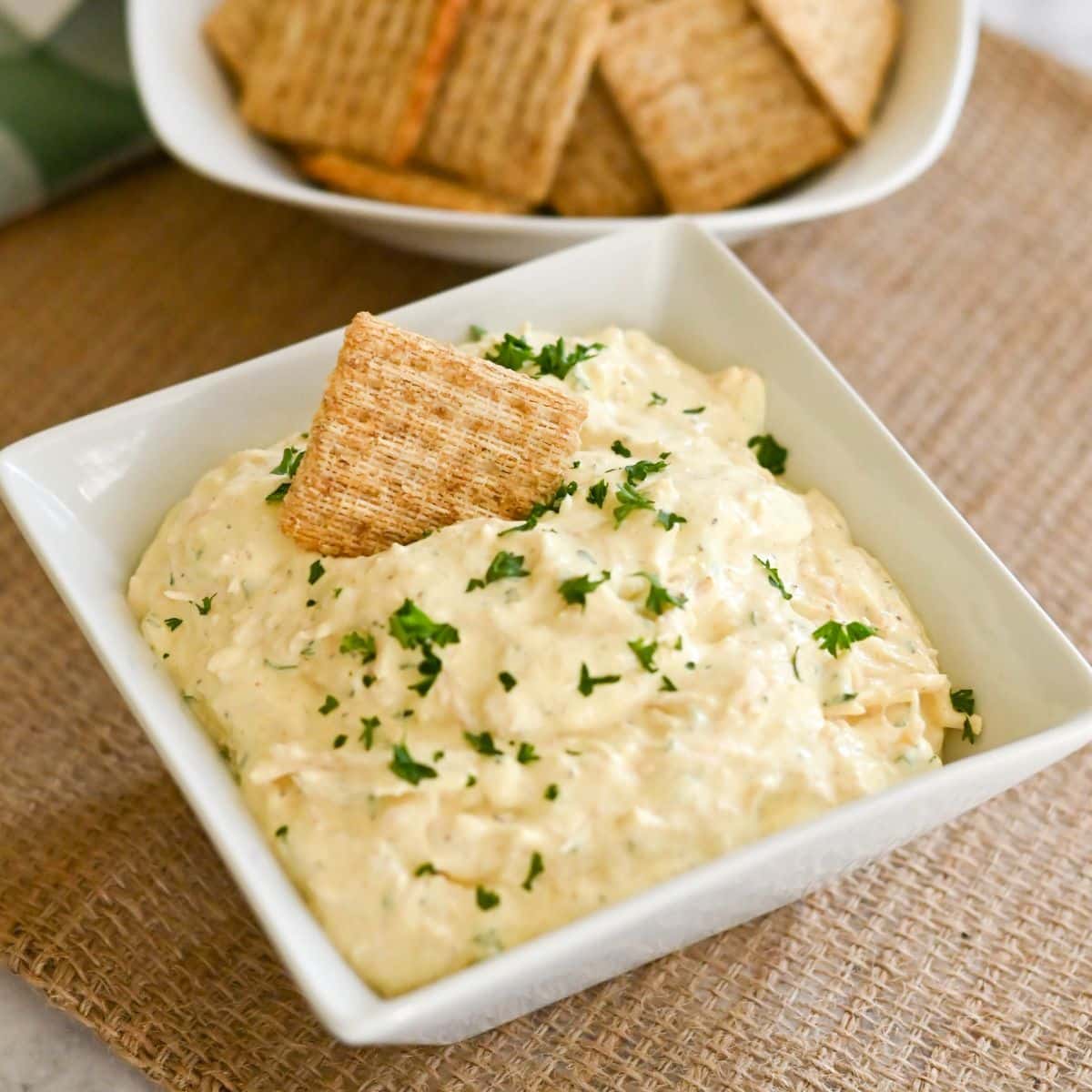 A square bowl of creamy chicken dip topped with parsley sits invitingly, surrounded by crispy crackers on a textured mat, with more crackers in the background.