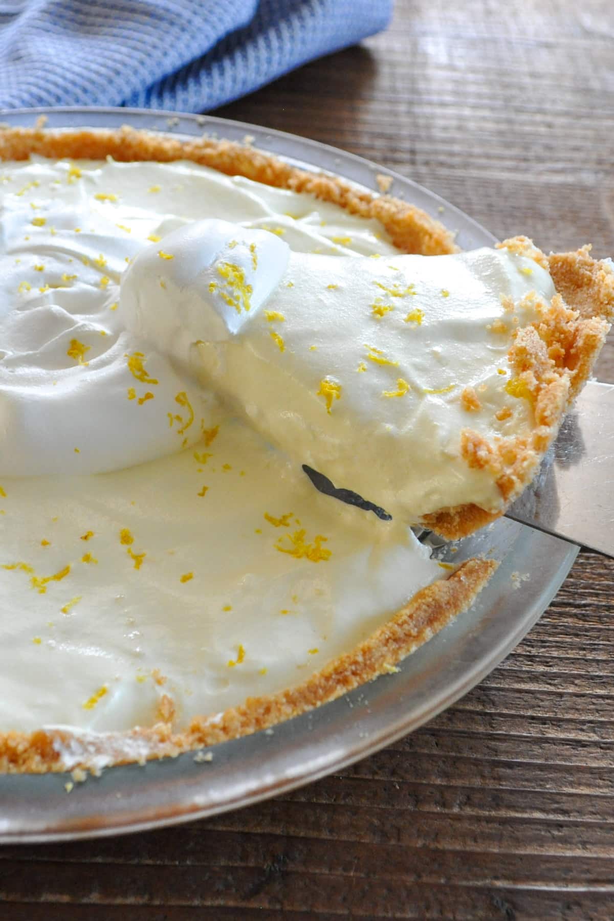 A creamy no-bake lemon pie is topped with whipped cream and lemon zest, with a slice being served from a glass pie dish.