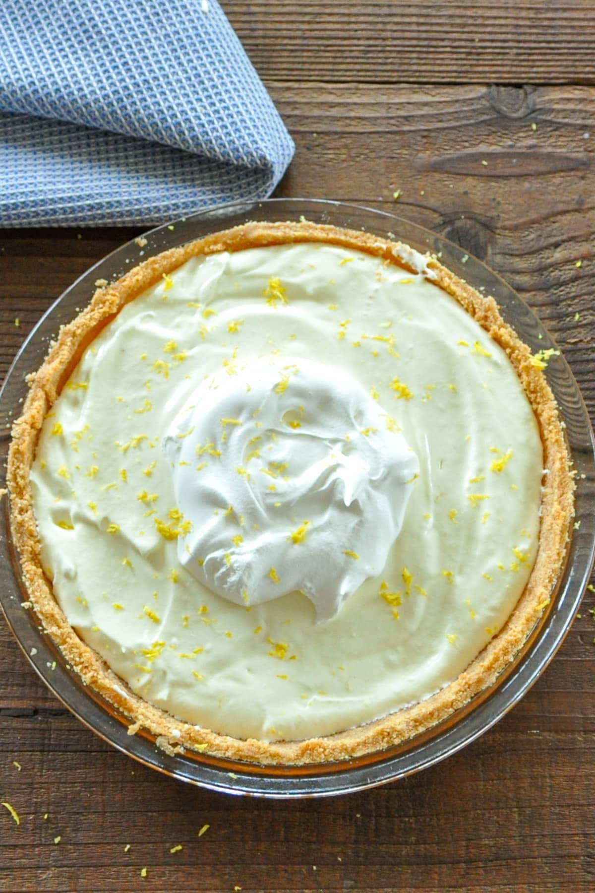 A creamy Lemon Marshmallow Pie topped with whipped cream and lemon zest sits in a glass dish on a wooden table, accented by a nearby blue cloth.