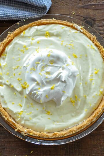 A no-bake lemon pie with whipped cream and lemon zest graces a glass dish on a wooden table, nestled near a blue cloth.
