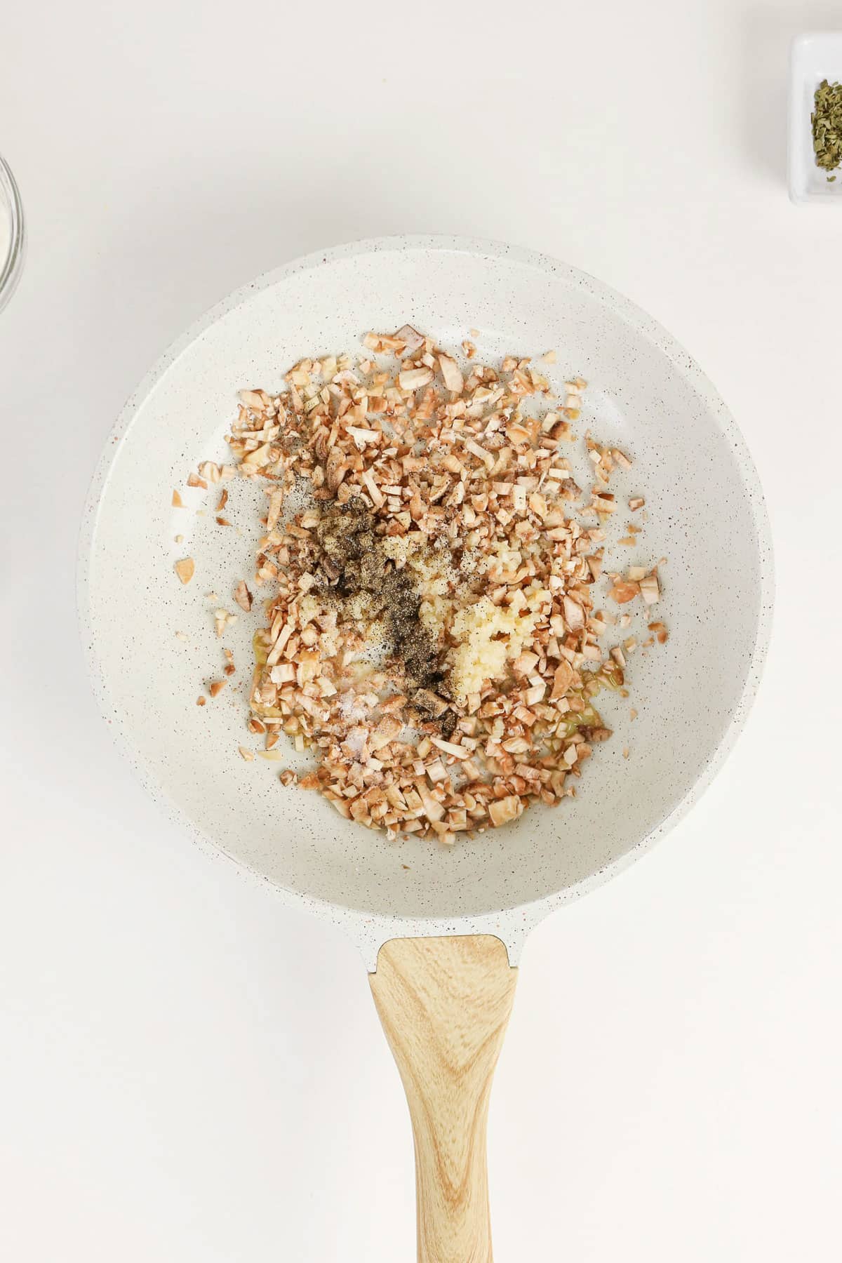 Chopped mushrooms and seasonings simmer in a white frying pan with a wooden handle on a light background, perfect for preparing Easy Stuffed Mushrooms.