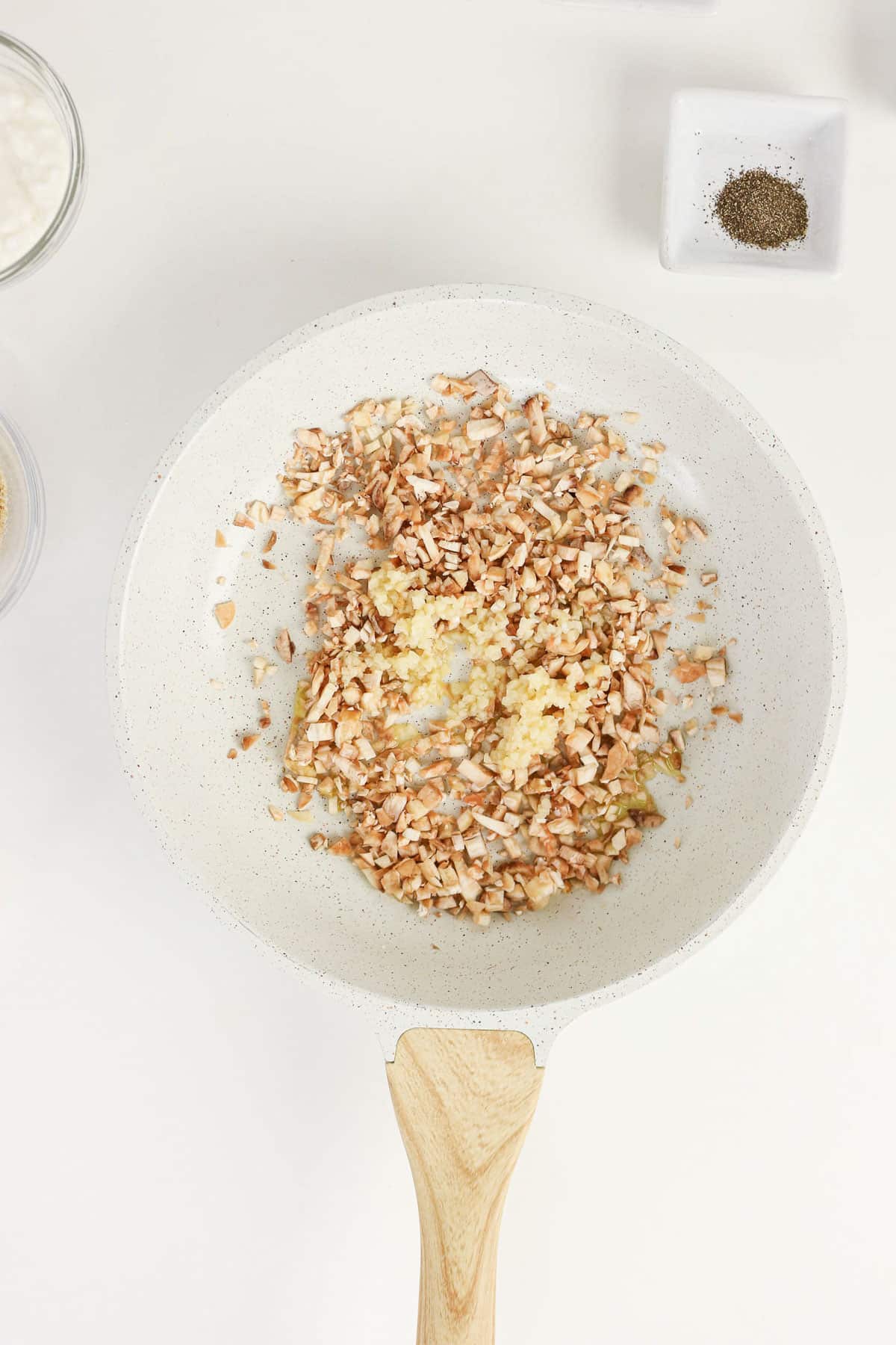 Chopped mushrooms and minced garlic, the perfect start for Easy Stuffed Mushrooms, cooking in a white skillet on a light countertop.