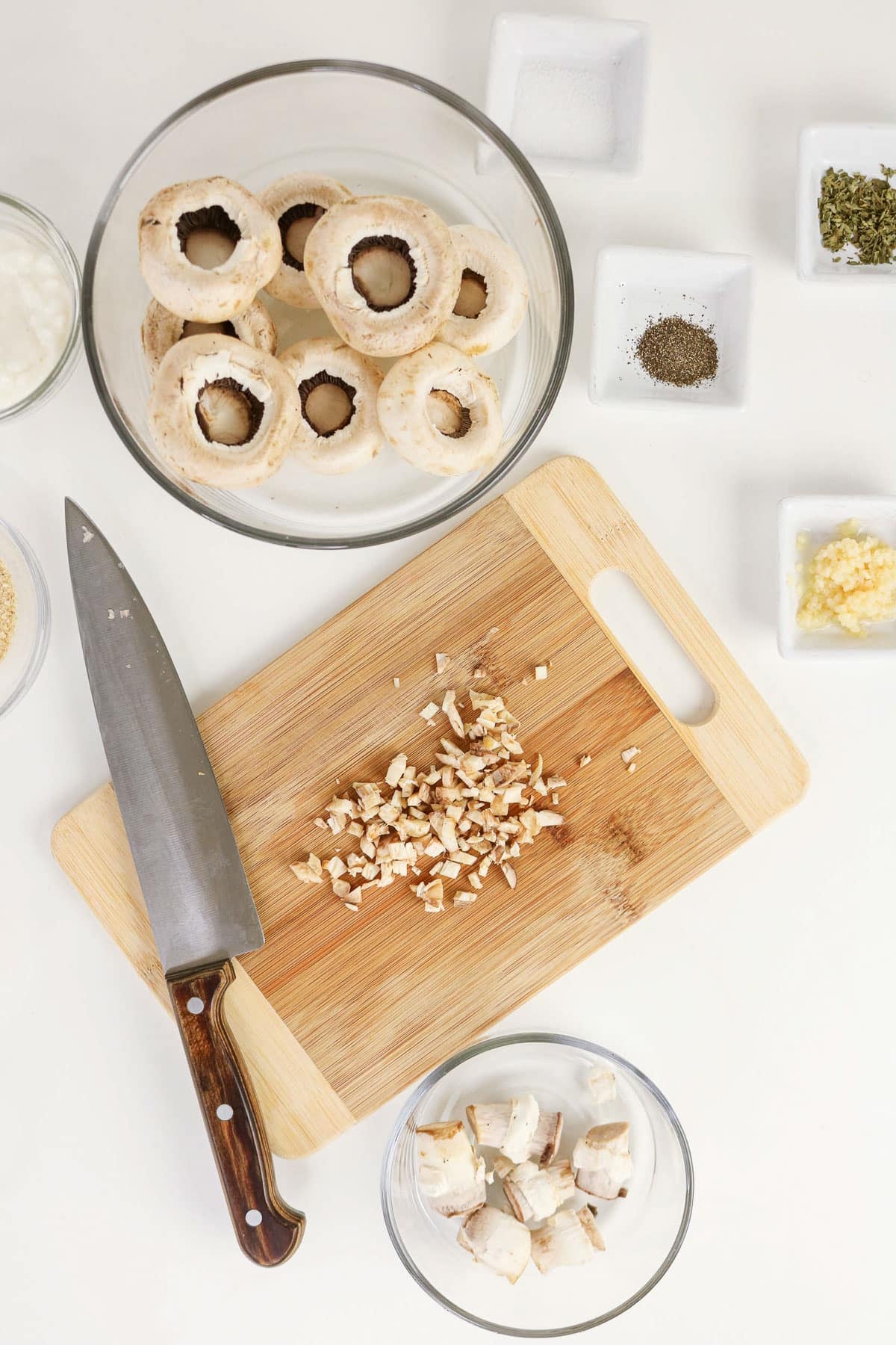 Chopped mushrooms on a cutting board with a knife, whole mushrooms, and seasonings on a white surface-perfect ingredients for Easy Stuffed Mushrooms.
