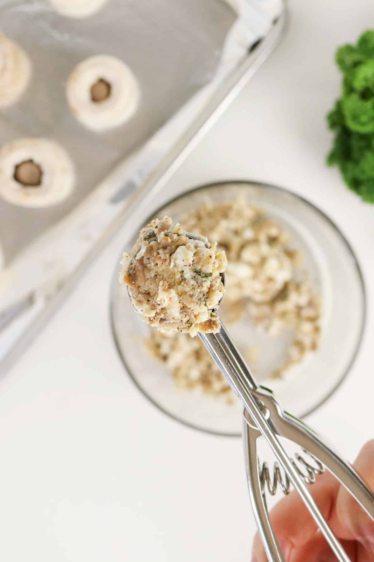 A cookie scoop holds herbed cheese filling above a bowl, ready for Easy Stuffed Mushrooms, with mushrooms and parsley nearby on a white surface.