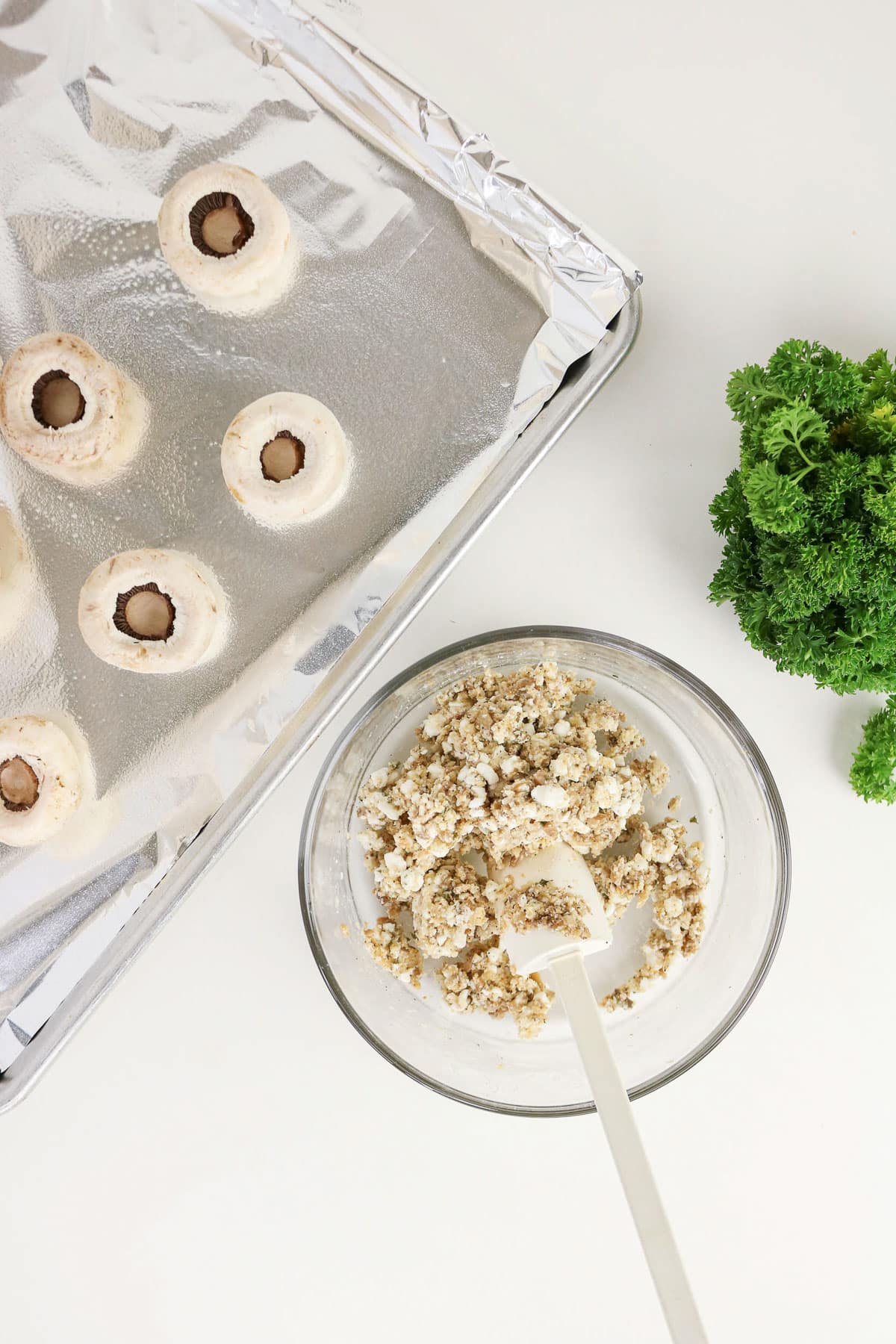 A bowl of stuffing, hollow mushrooms ready for filling on a foil-lined tray, and fresh parsley hint at delicious Easy Stuffed Mushrooms soon to be enjoyed.