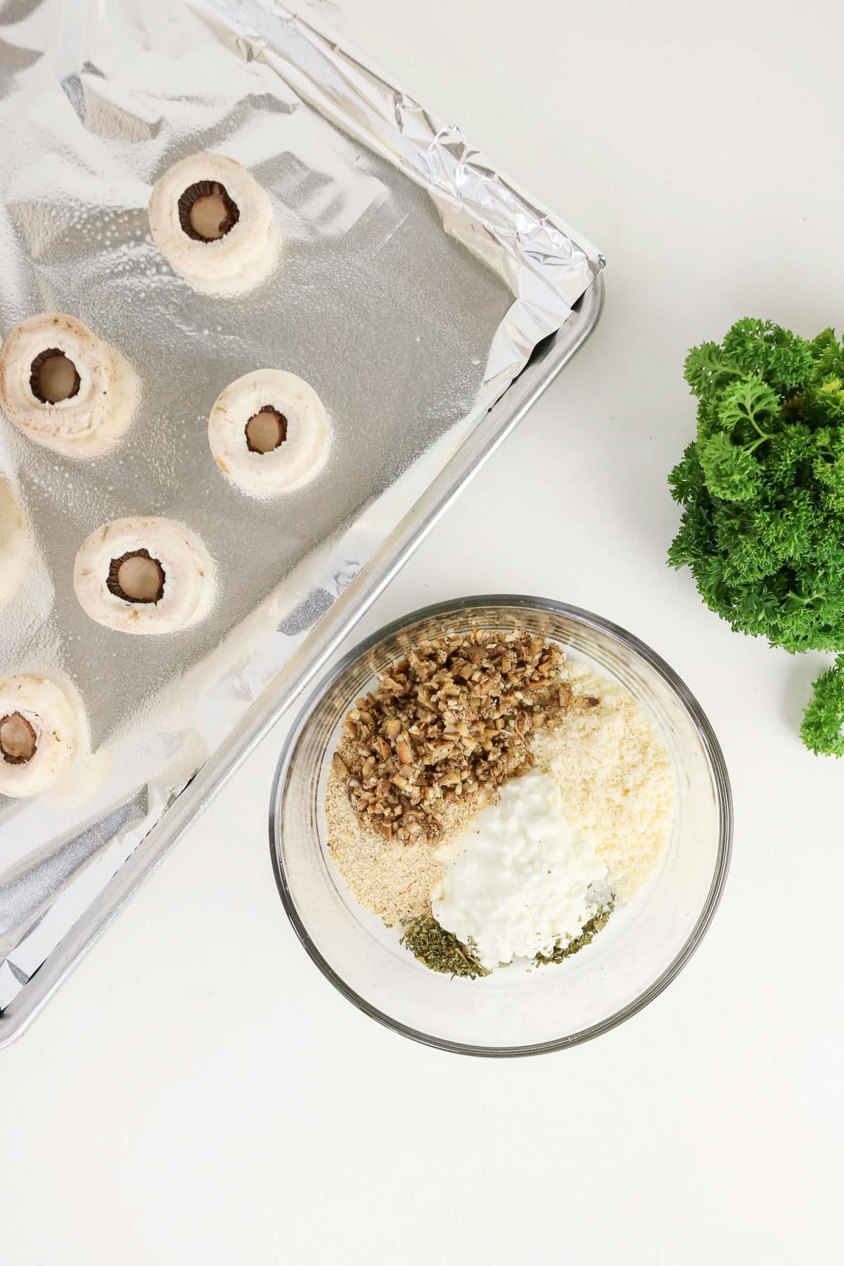 A bowl with Easy Stuffed Mushrooms ingredients, a foil-lined tray with hollow mushrooms, and fresh parsley on a white table.