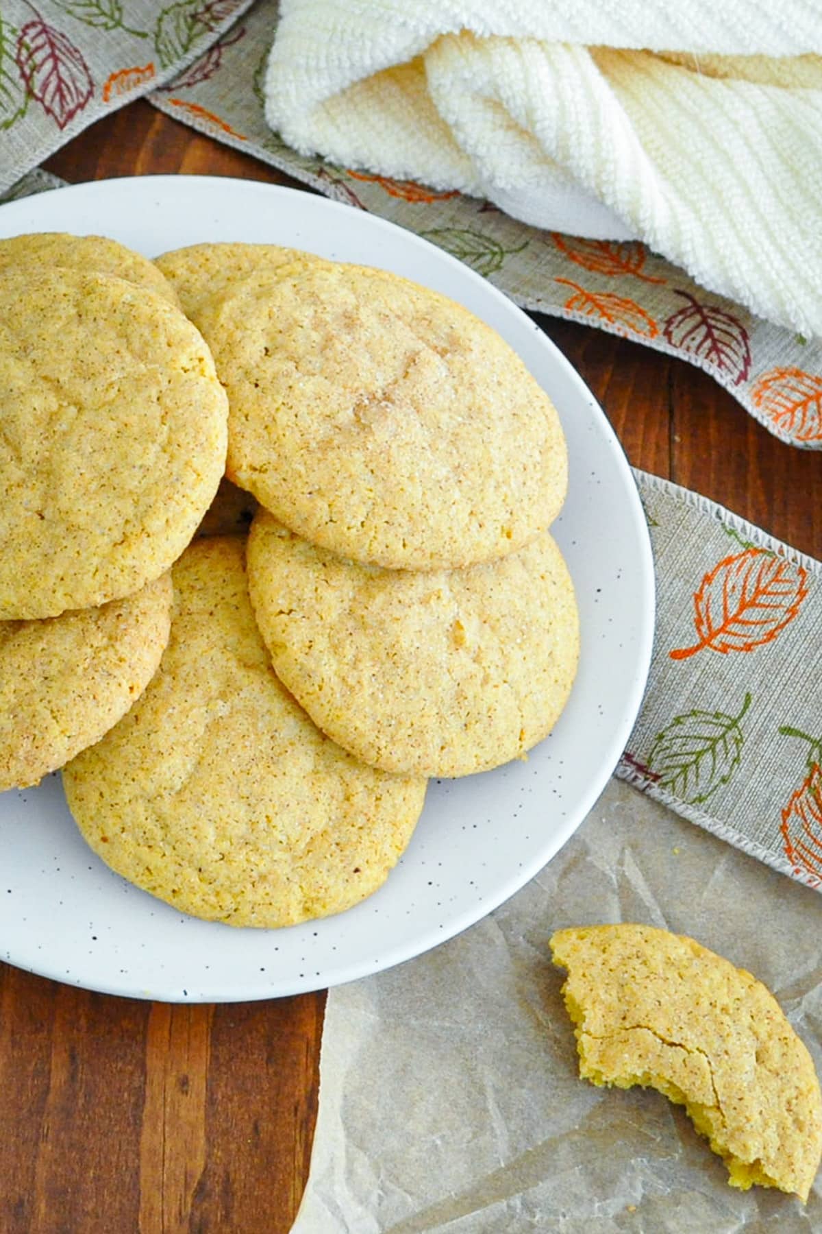 Overhead view of pumpkin snickerdoodles on a white plate, with a bitten cookie on parchment beside it.