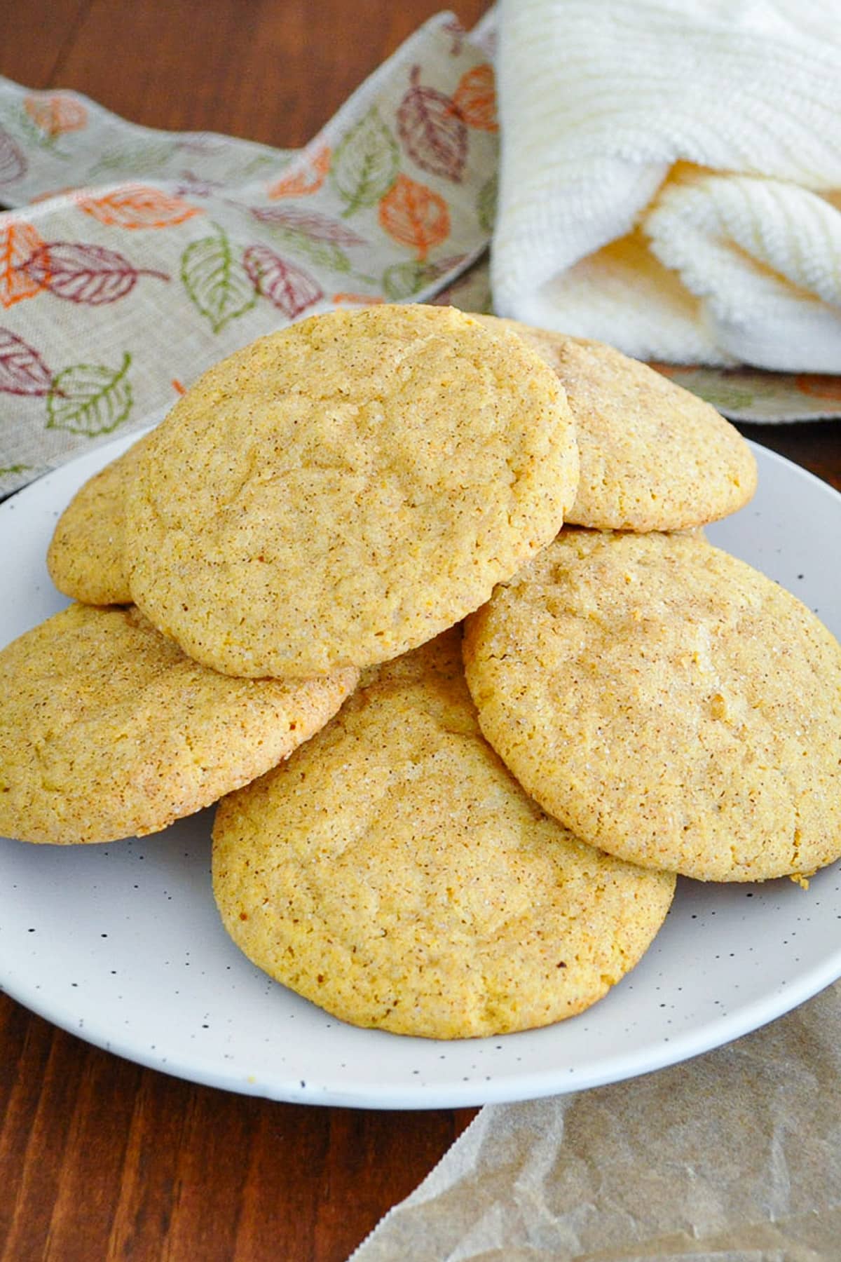 A pile of pumpkin snickerdoodles on a speckled white plate, with a fall leaf ribbon and cream towel behind it.