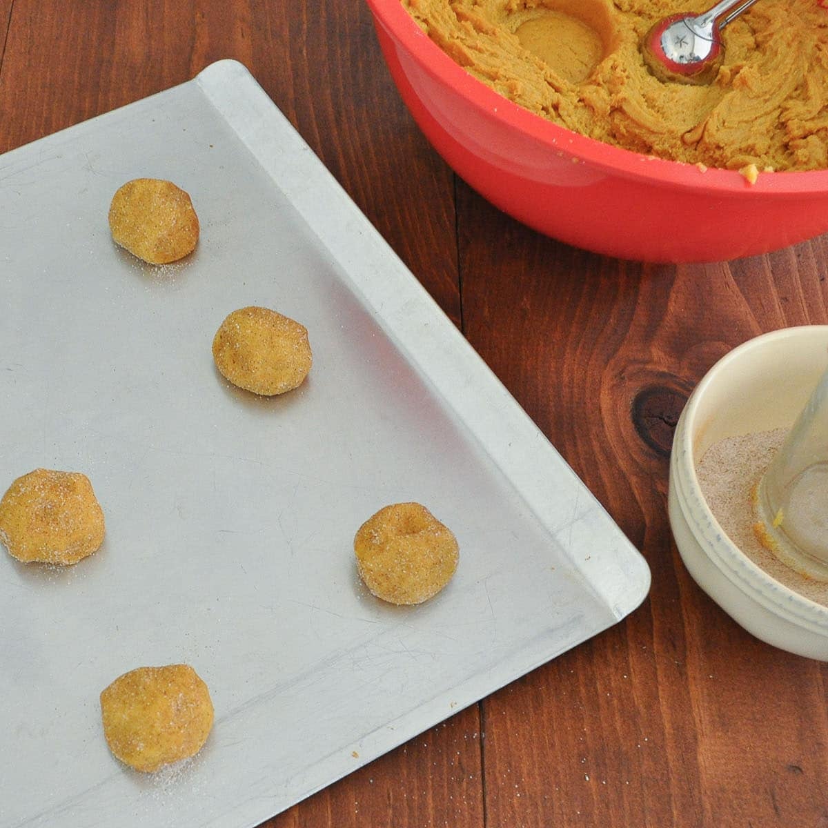 Sugar-coated dough balls on a cookie sheet next to the bowl of dough and a glass resting in a bowl of spiced sugar.