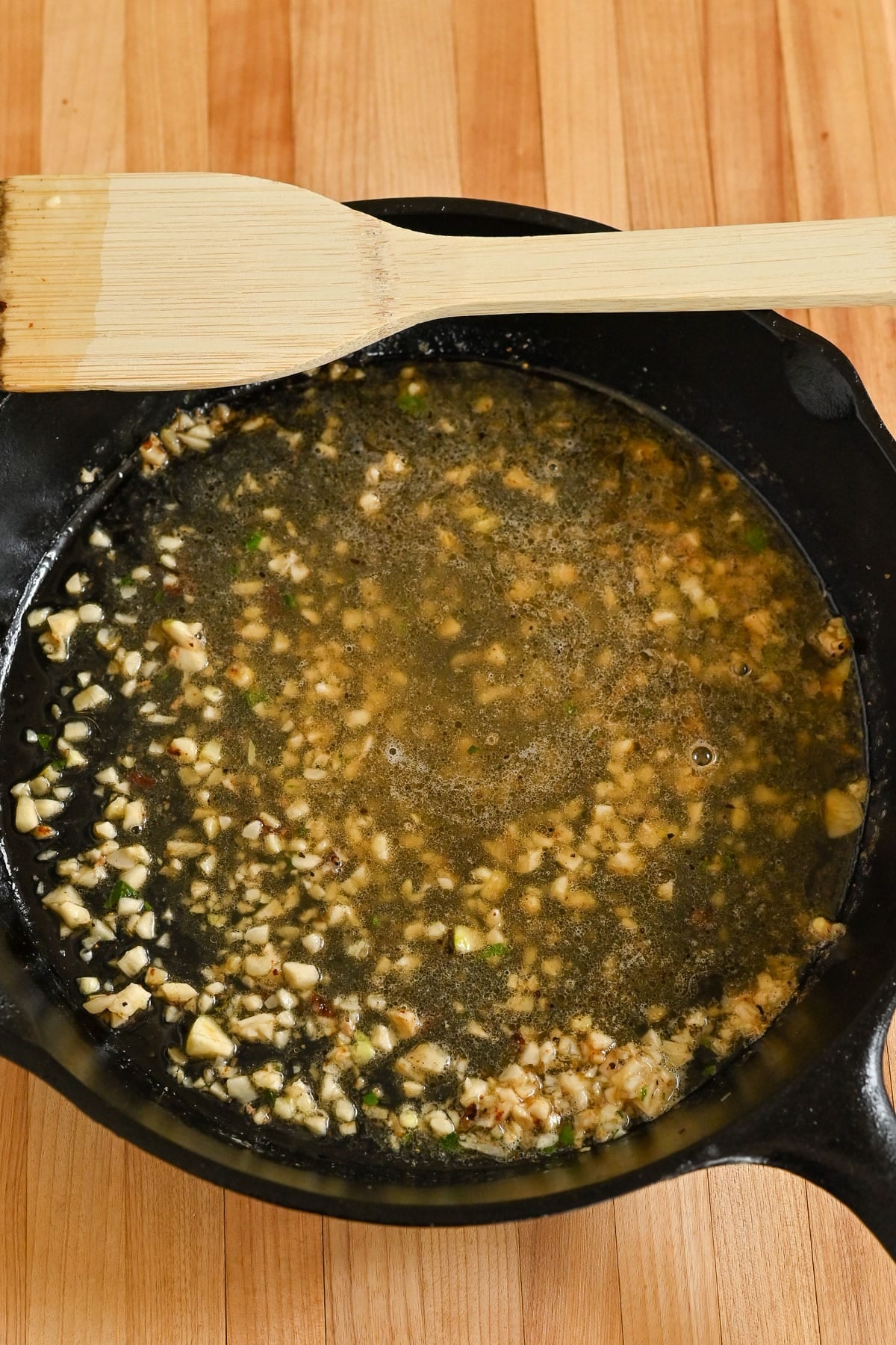 Chopped garlic cooking in a skillet with oil, perfect for making a flavorful garlic sauce, next to a wooden spatula on a wooden countertop.