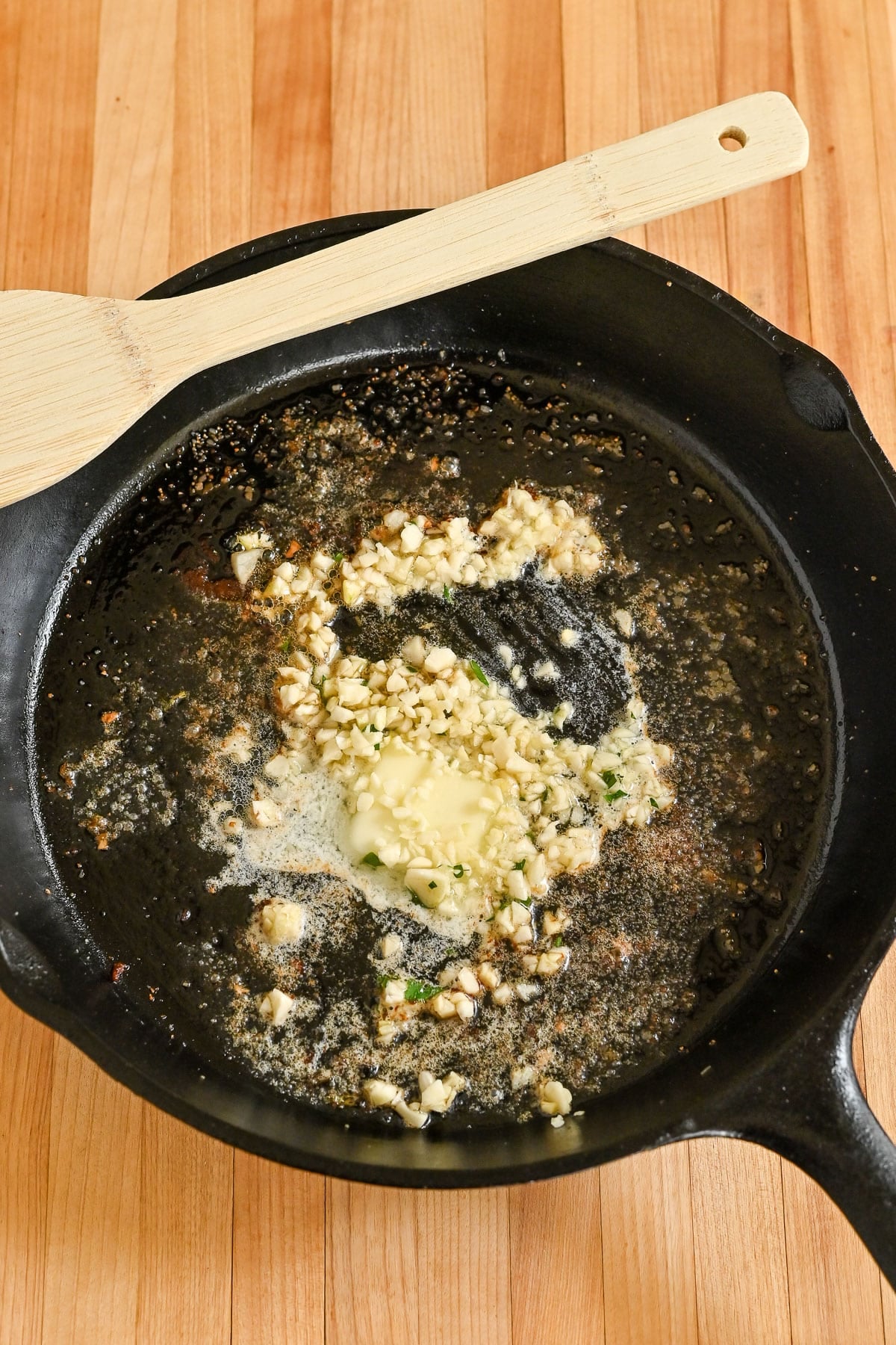 Chopped garlic and melting butter sizzling in a black cast iron skillet, the perfect start for Pork Chops with Garlic White Wine Sauce, on a wooden surface with a spatula.