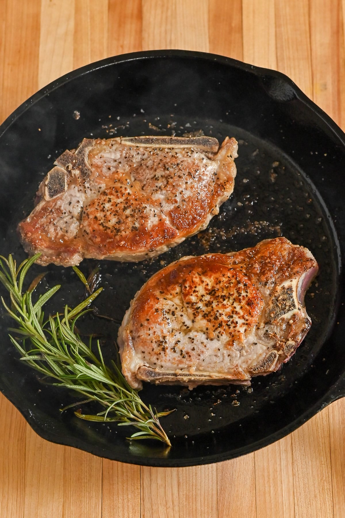 Two cooked Pork Chops with Garlic White Wine Sauce in a cast iron skillet, garnished with pepper and a sprig of fresh rosemary.