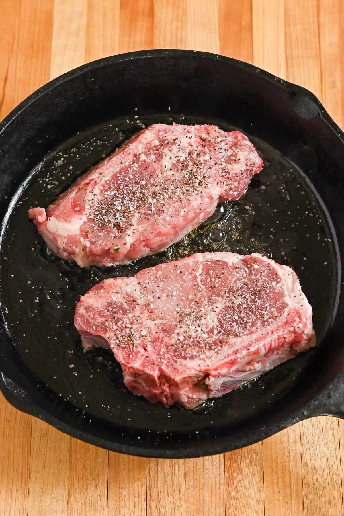 Two raw, seasoned steaks in a black cast iron skillet on a wooden surface, ready to be transformed with a rich garlic sauce or white wine sauce.
