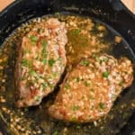 Two cooked steaks in a skillet topped with garlic, herbs, and a savory white wine sauce.