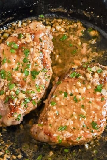 Two cooked steaks in a skillet topped with garlic, herbs, and a savory white wine sauce.