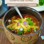 A mug of hearty Ground Turkey Chipotle Chili topped with melty cheese, green onions, and chopped red onions sits invitingly on a table.