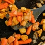 A spatula lifts roasted carrots with apples and onions, all seasoned with herbs on a dark surface.