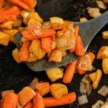 A spatula lifts roasted carrots with apples and onions, all seasoned with herbs on a dark surface.