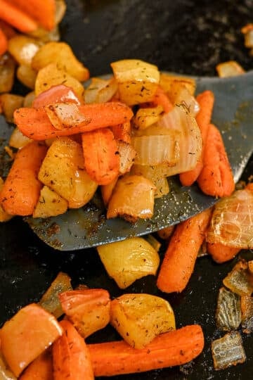 A spatula lifts roasted carrots with apples and onions, all seasoned with herbs on a dark surface.