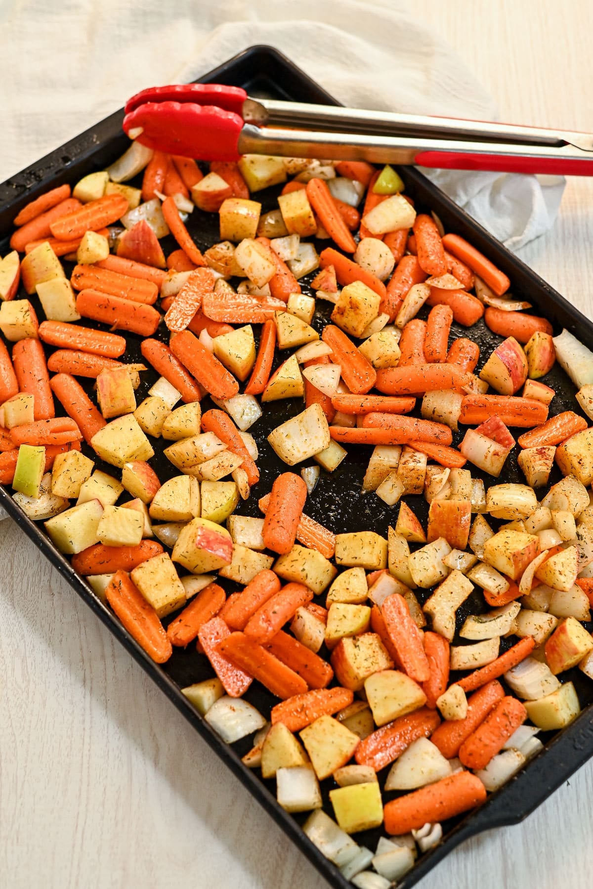 Sheet pan with roasted carrots with apples and onions, plus potatoes, red tongs placed on the side.