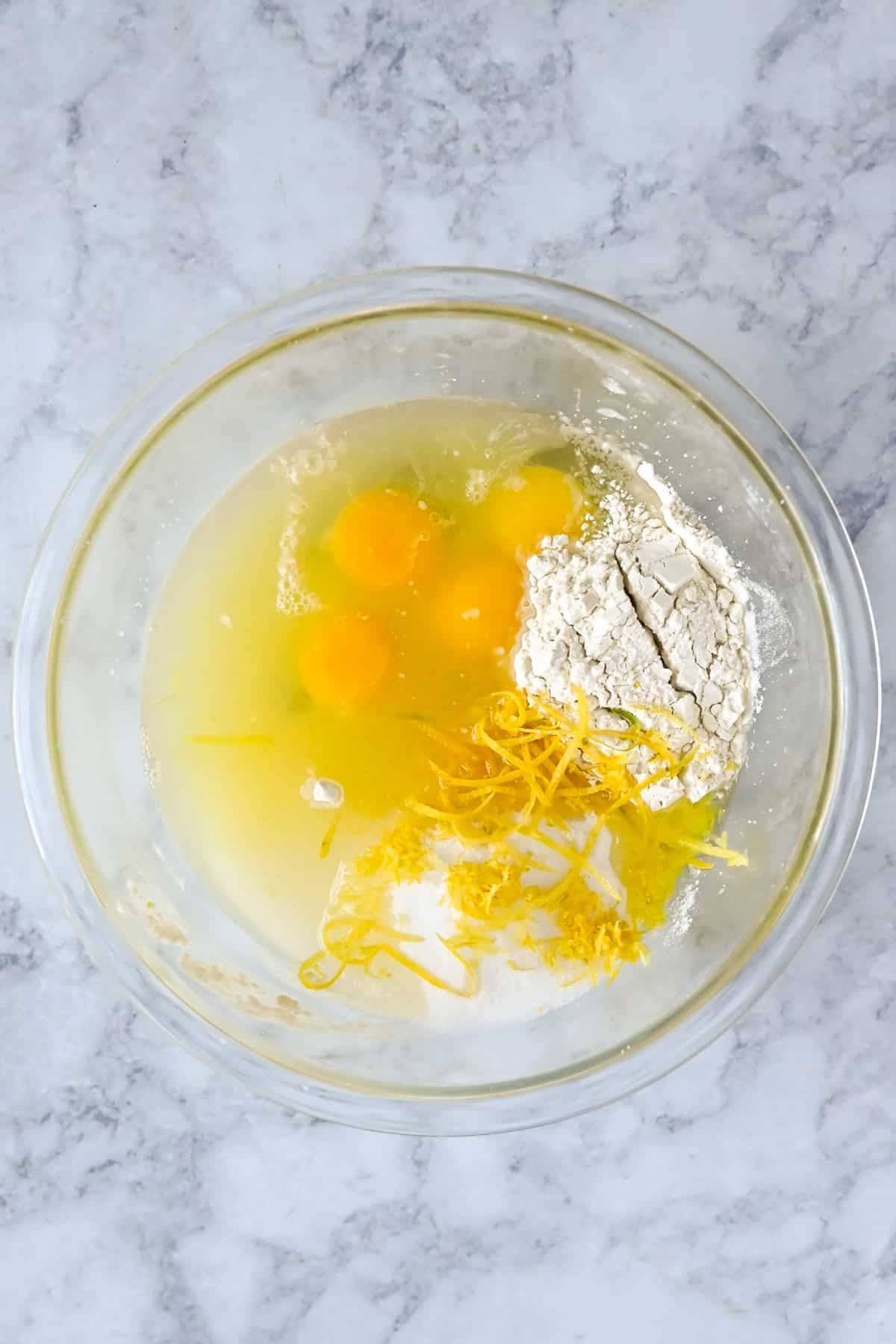 A glass bowl with eggs, flour, sugar, lemon zest, and liquid on a marble surface, ready for mixing-perfect ingredients for baking tart lemon bars or delicious lemon cookies.