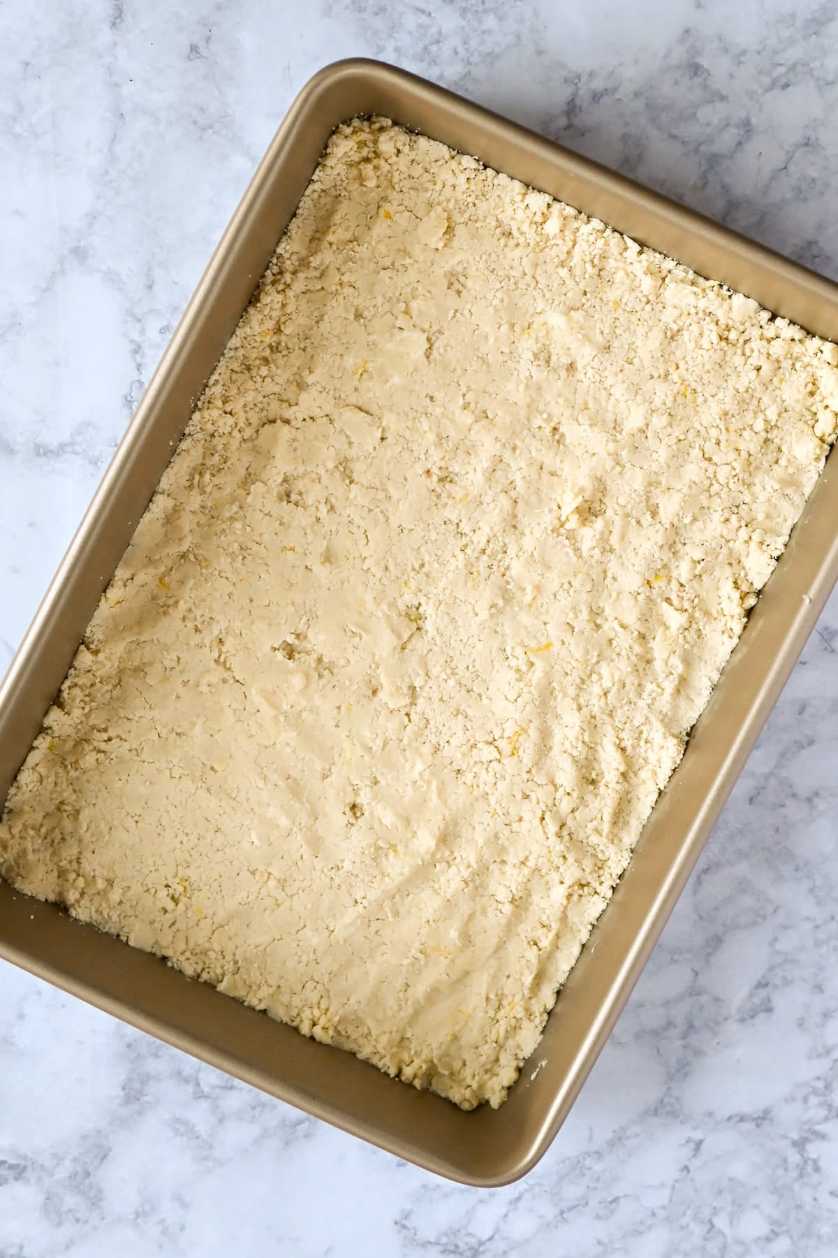 Unbaked crumbly dough for Tart Lemon Bars is pressed evenly into a rectangular metal baking pan on a marble surface.