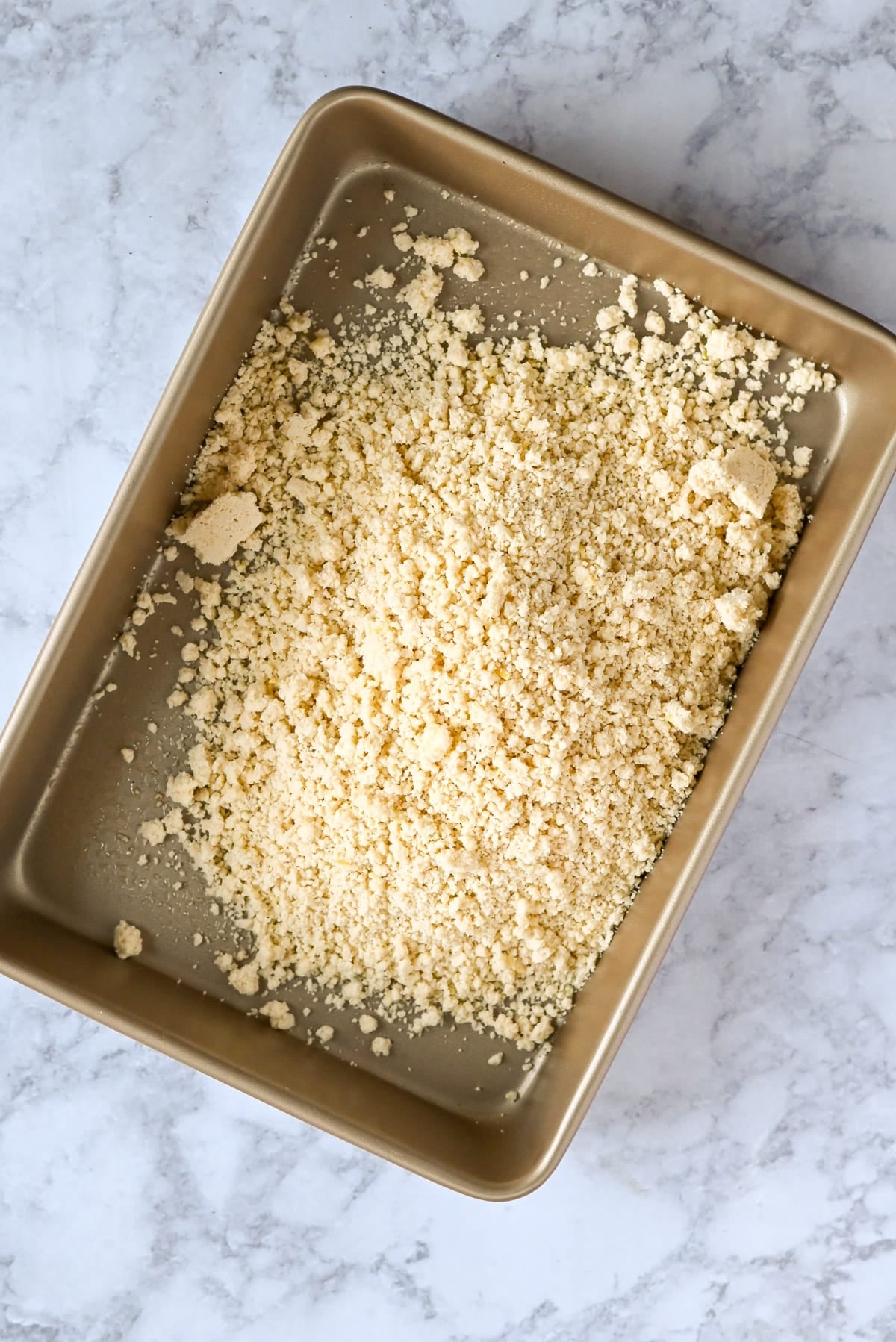 A gold baking pan filled with crumbly, pale yellow streusel topping-evoking the bright, zesty appeal of Tart Lemon Bars-rests on a marble surface.
