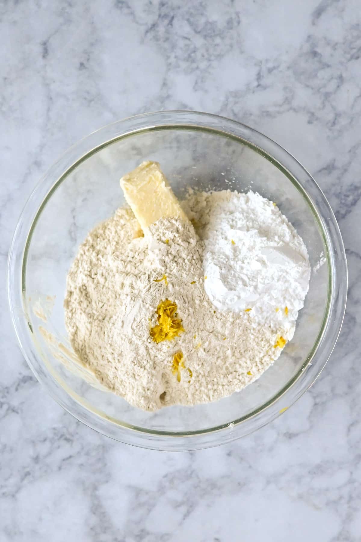 Glass bowl with flour, butter, powdered sugar, and lemon zest on a marble countertop-perfect for preparing Tart Lemon Bars or zesty lemon cookies.