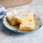 Four Tart Lemon Bars dusted with powdered sugar on a blue plate with a decorative paper doily.