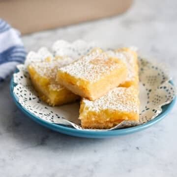 Four Tart Lemon Bars dusted with powdered sugar on a blue plate with a decorative paper doily.