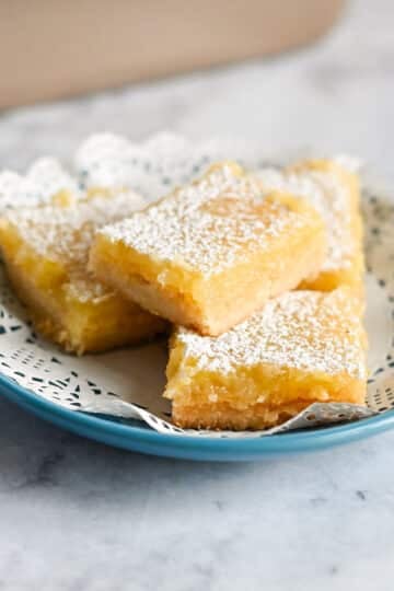 Four Tart Lemon Bars dusted with powdered sugar on a blue plate with a decorative paper doily.